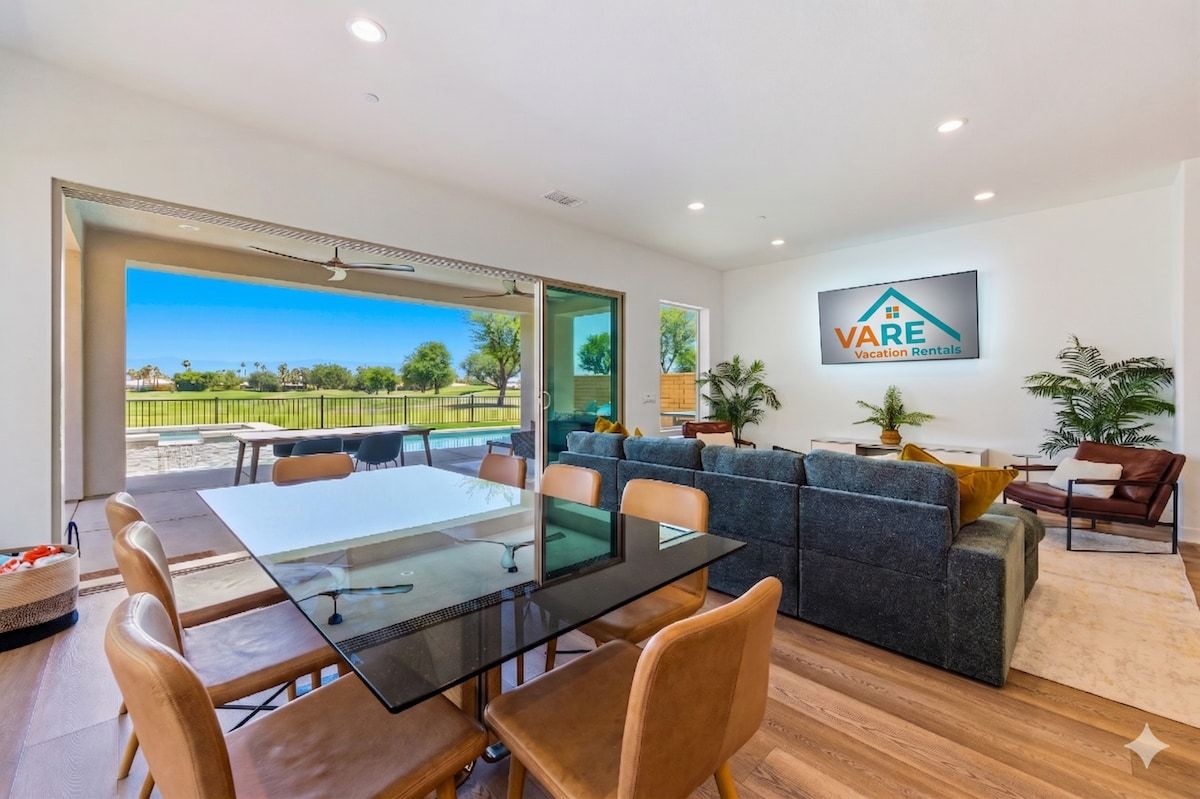 A modern living room with a glass table and chairs, a grey sofa, and a VARE Vacation Rentals logo on the wall.