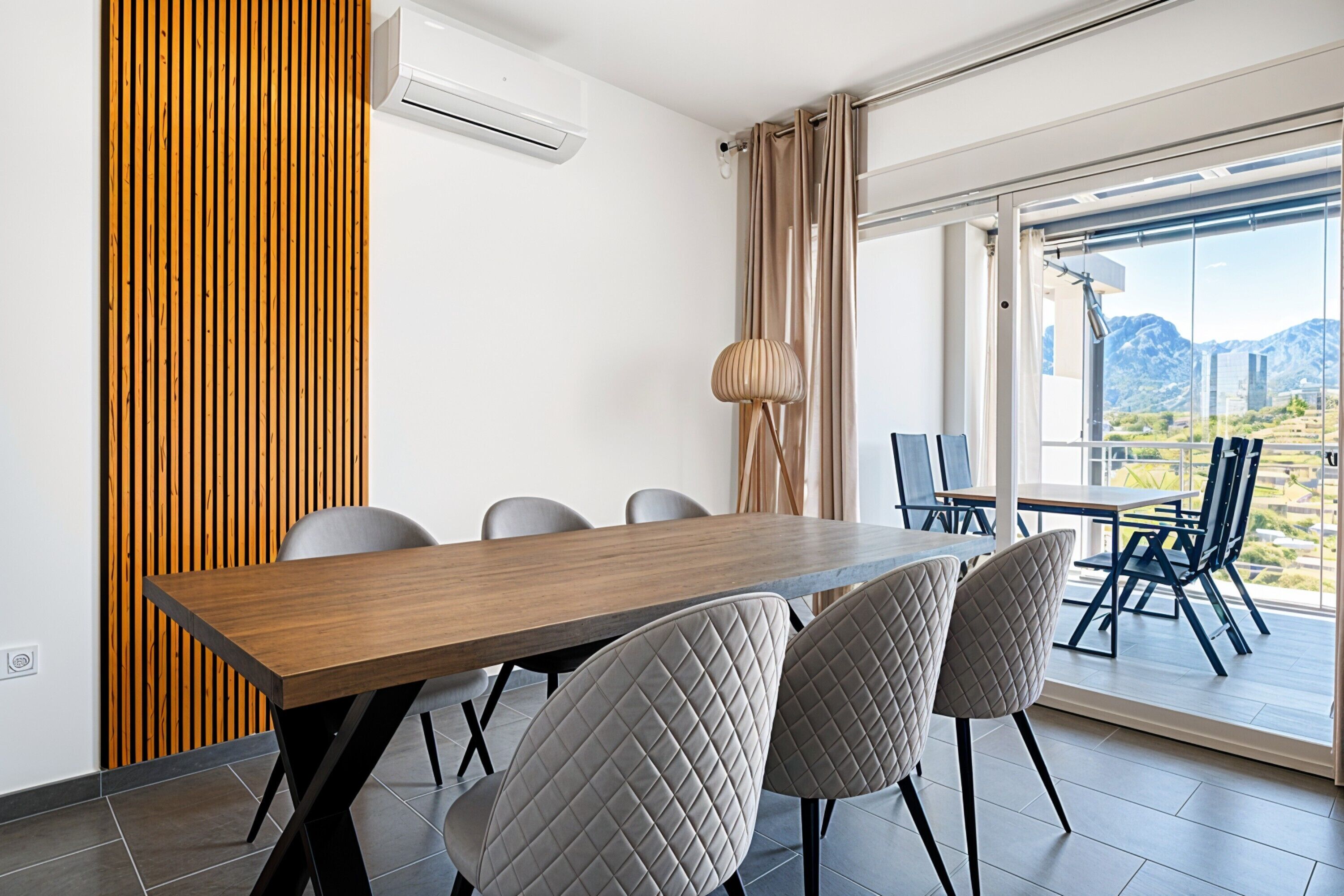 A modern dining area with a wooden table, four chairs, and a view of mountains through large windows.