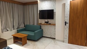 Una stanza con un divano color petrolio, un tavolino in legno, una TV montata su una parete con effetto marmo, una porta con maniglia e una finestra con tende.