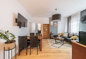 A modern living room with a dining area, a large wooden bookshelf, a TV, and a clock on the wall. - RentPlanet - Apartament Bukowska (Poznań)