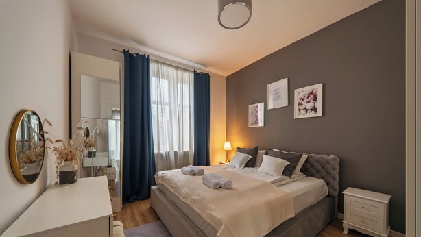 Un dormitorio con cama, mesitas de noche, cómoda, espejo y ventana con cortinas.