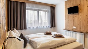 Een slaapkamer met een bed, twee kussens, een tv bevestigd aan een houten muur, en een raam met gordijnen.