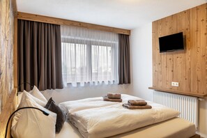 Une chambre à coucher avec un lit, deux oreillers, un téléviseur fixé au mur (qui est en bois) et une fenêtre avec des rideaux.