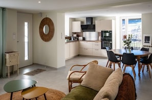 Eine moderne Küche mit weißen Schränken, ein Essbereich mit Holztisch und -stühlen sowie ein Wohnbereich mit Sofa und Couchtisch.