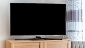 Un téléviseur posé sur un meuble en bois, avec une télécommande à proximité, un rideau à motifs et un canapé gris dans une pièce.