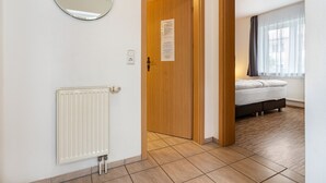 Une pièce avec une porte en bois, un radiateur blanc, un lit avec un matelas blanc et une fenêtre avec des rideaux.