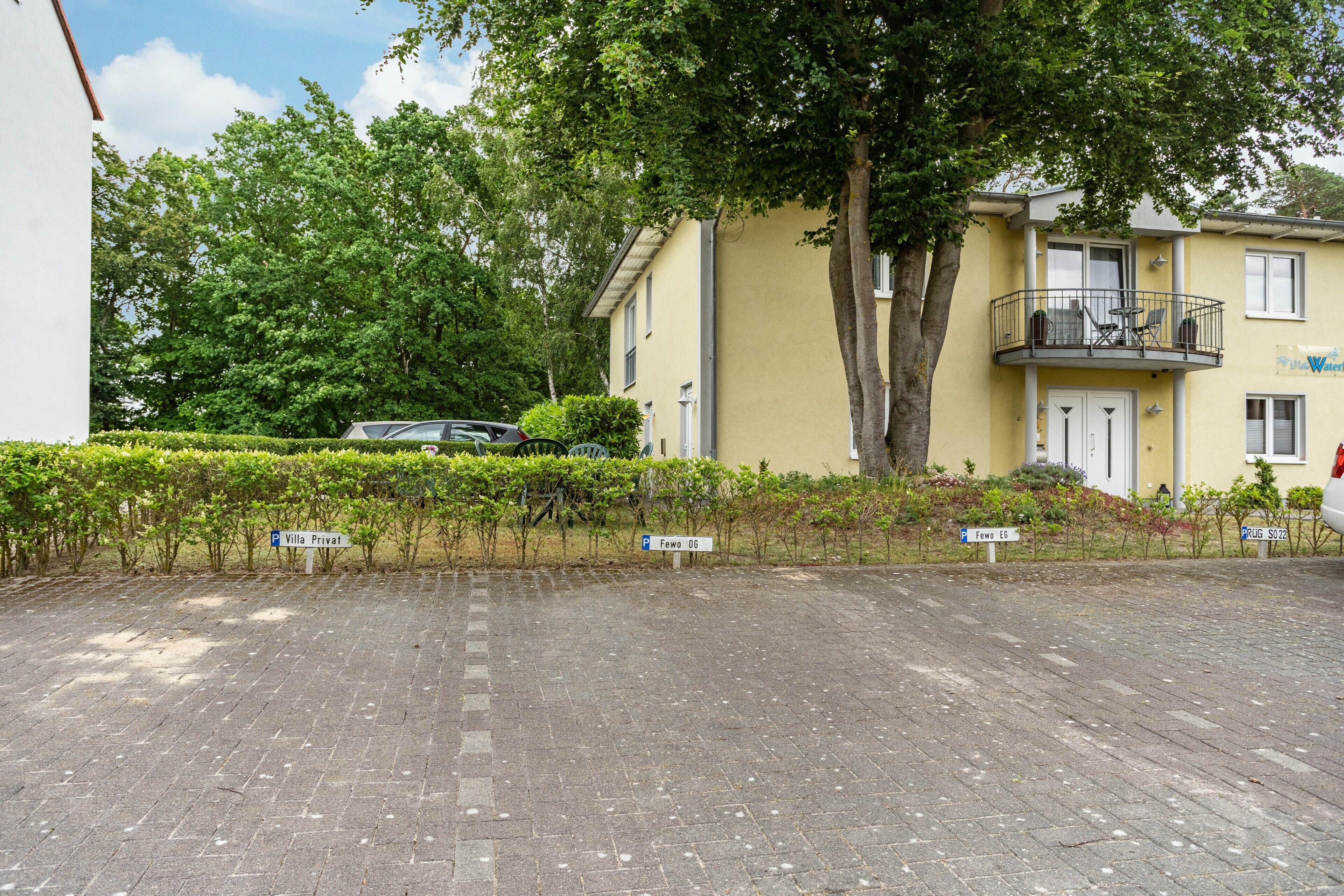 Uma área de estacionamento pavimentada com um edifício amarelo, uma árvore e uma placa com a inscrição 'Villa Privat'.