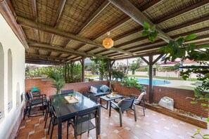 Um pátio externo com mesa e cadeiras, um lustre pendurado e vista para uma piscina.