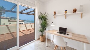 Een balkon met uitzicht op zee, een bureau met een laptop, een bloempot en decoratieve items op planken.