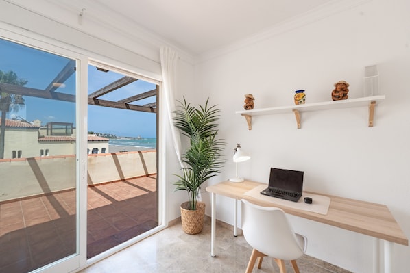 A balcony with a view of the sea, a desk with a laptop, a potted plant, and decorative items on shelves.