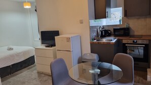 Une chambre à coucher de taille modeste, comprenant un lit, une petite commode blanche, un téléviseur et une table en verre avec deux chaises.