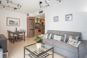 Un salon moderne avec une table basse en verre, un canapé gris agrémenté de coussins colorés, un coin repas avec une table et des chaises, et une cuisine en arrière-plan.