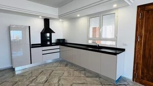 A kitchen with a black range hood, white cabinets, a black countertop, and a window with blinds. - Maisons Jassim (Essouani)