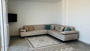 A living room with a beige sectional sofa, a patterned rug, and a flat-screen TV mounted on the wall. - Maisons Jassim (Essouani)