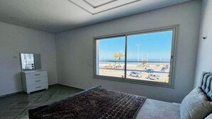 A bedroom with a bed, a dresser, a window with a view of the beach and parking lot. - Maisons Jassim (Essouani)