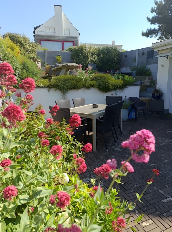 Eine Terrasse mit Tisch und Stühlen, umgeben von üppigem Grün und rosa Blüten.