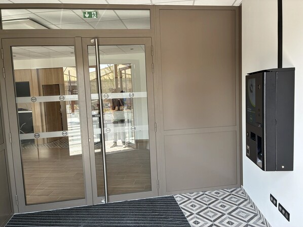 A hotel entrance with a glass door, a patterned rug, and a wall-mounted device. - B&B Hotel Amiens Sud (Amiens)