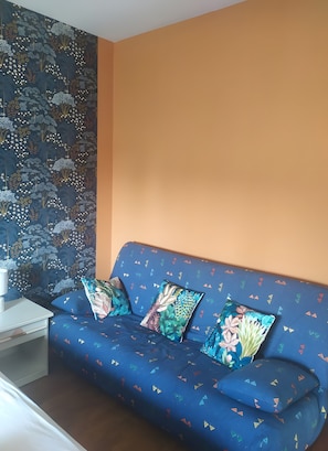 Een kamer met een blauwe bank tegen een oranje muur, een patroonrijk wandtapijt en een witte bijzettafel.