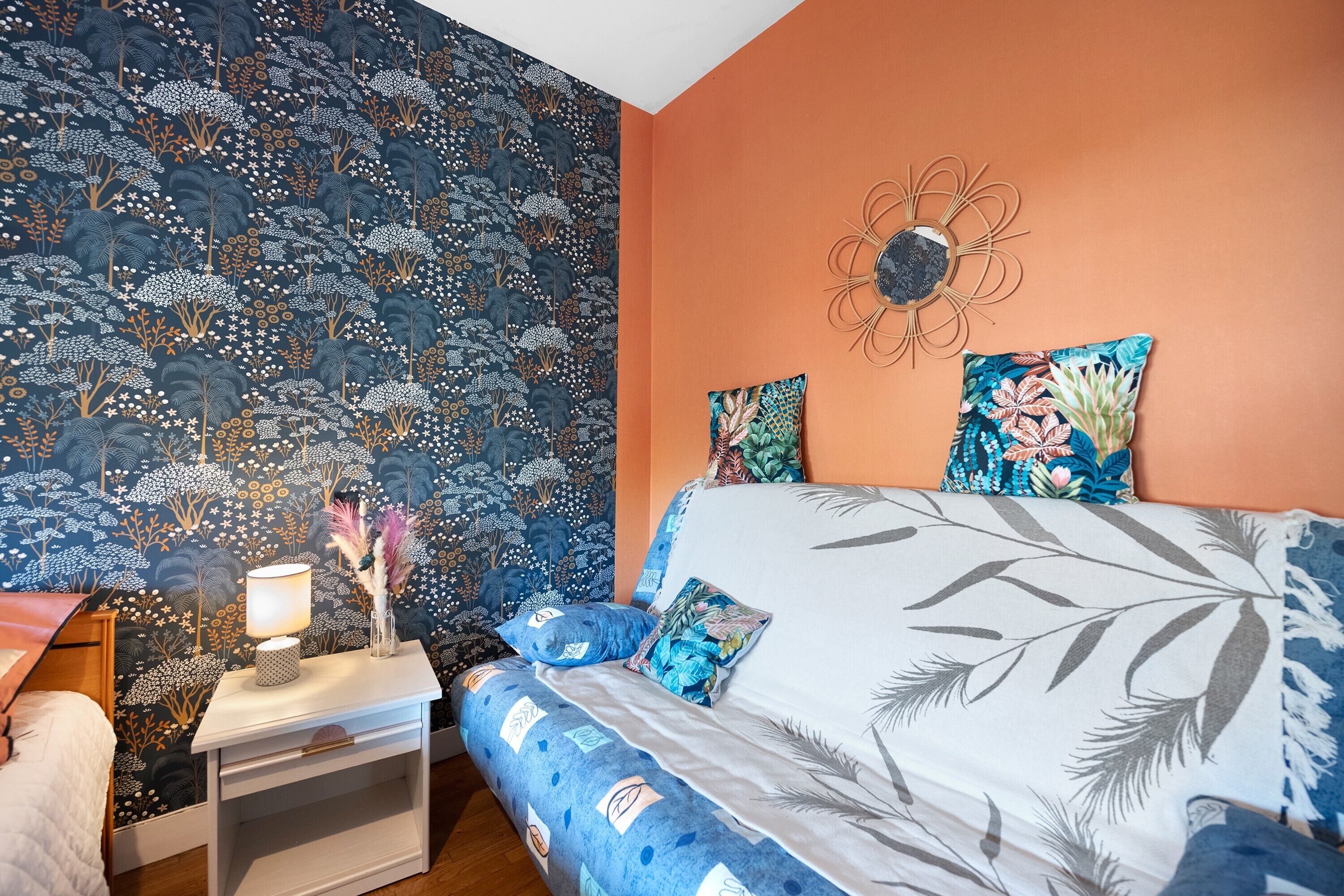 A bedroom with a patterned wallpaper, a bed with floral pillows, a nightstand with a lamp, and a decorative wall piece.