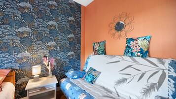 A bedroom with a patterned wallpaper, a bed with floral pillows, a nightstand with a lamp, and a decorative wall piece.
