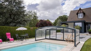 Una piscina con cerramiento de vidrio, sillas rosas y una casa al fondo.