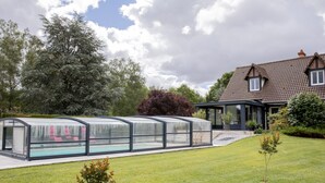A house with a swimming pool covered by a glass structure, a garden with small trees, and a large tree in the background.