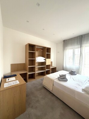 Ein Schlafzimmer mit einem Bett, einem Holzschrank, einem Schreibtisch mit Papieren und einem Fenster mit Vorhängen.