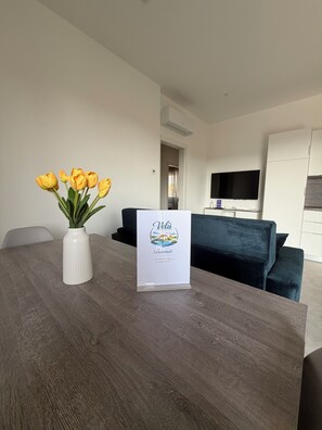 Ein modernes Wohnzimmer mit einem Holzesstisch, einer Vase voller gelber Tulpen, einem Willkommensschild, einem dunkelblauen Sofa und einem flachbildfernseher an der Wand.