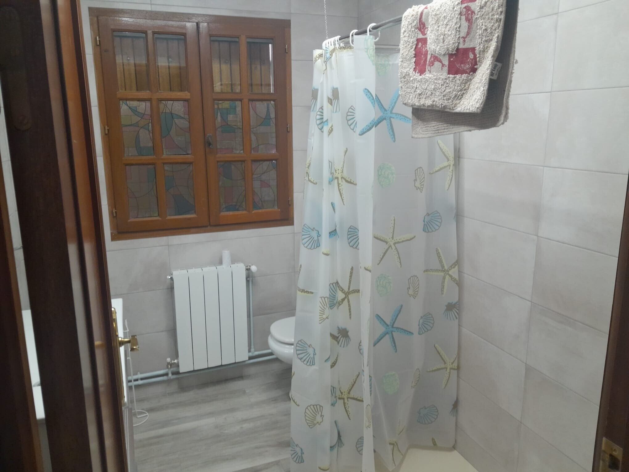 Un baño con cortina de ducha adornada con estrellas de mar y conchas, un radiador blanco, un ventanal de madera y una toalla colgada en la pared.