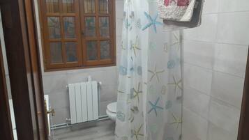 Un baño con cortina de ducha adornada con estrellas de mar y conchas, un radiador blanco, un ventanal de madera y una toalla colgada en la pared.