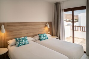A hotel room with a wooden headboard, two beds with white linens, two bedside lamps, and a balcony with a view of buildings and mountains. - Ereza Villas La Granja (Yaiza)