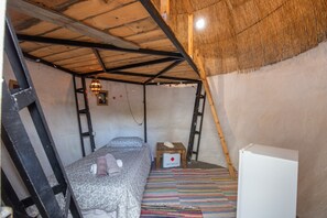 Een kleine, rustieke kamer met een hoogslaper, een klein tafeltje, een koelkast en een kleurrijk gestreept tapijt.