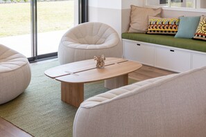 Uma sala de estar moderna com uma mesa de centro redonda de madeira, duas poltronas bege, um sofá com almofadas estampadas e vista através de grandes janelas.