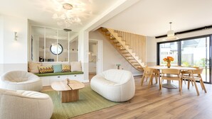 A modern living room with a staircase, a dining area, and a view through large windows.