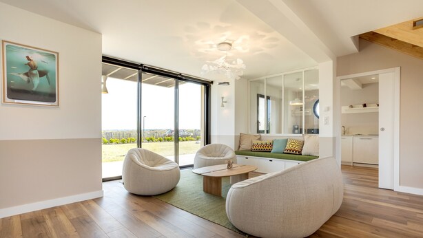 A modern living room with a large sliding glass door to the outdoors, a dining area with a round table, and a kitchen visible through an open door.