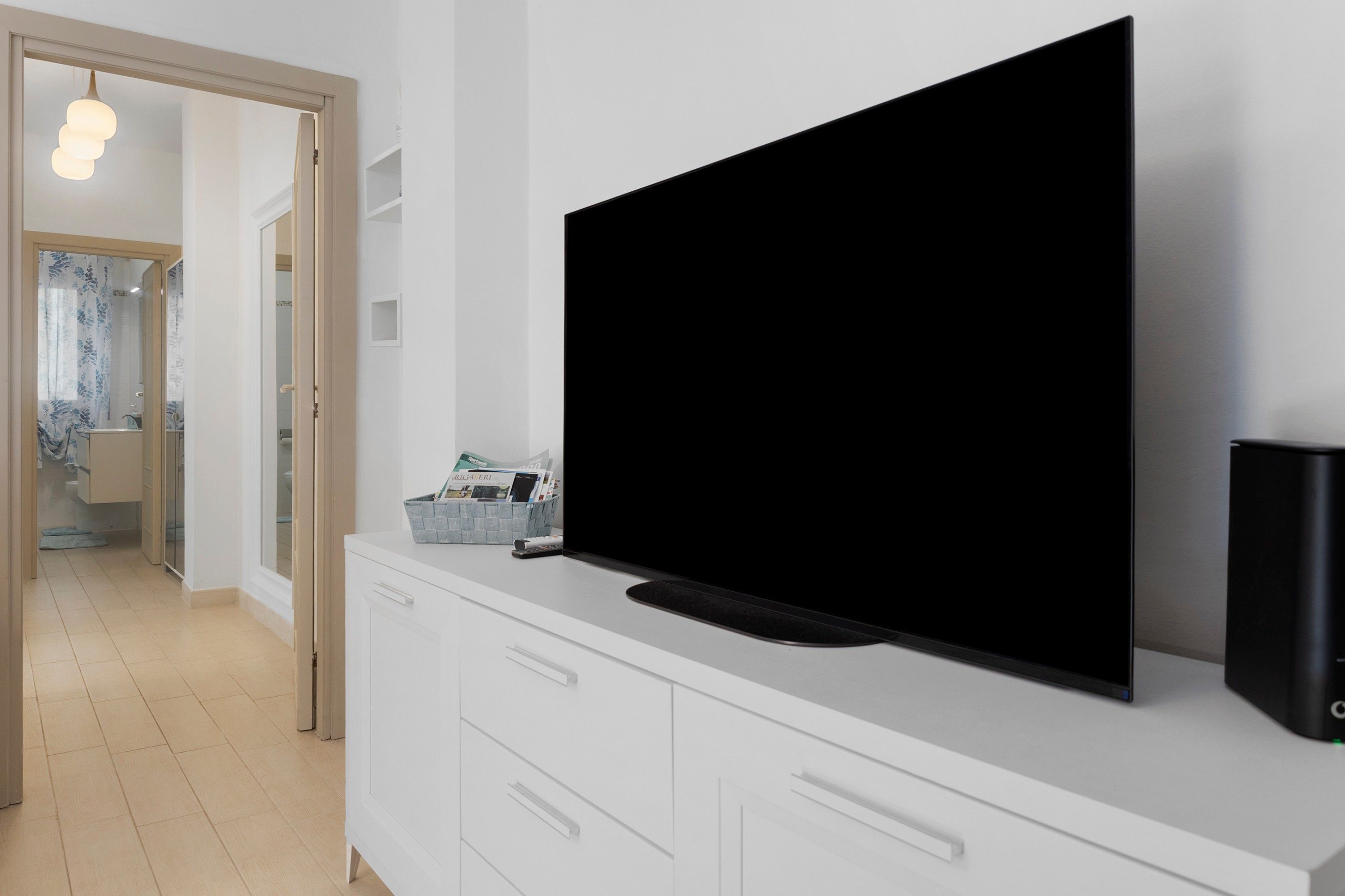 Un soggiorno moderno con un'ampia TV a schermo piatto montata su un mobile bianco, un telecomando e un altoparlante sulla destra.