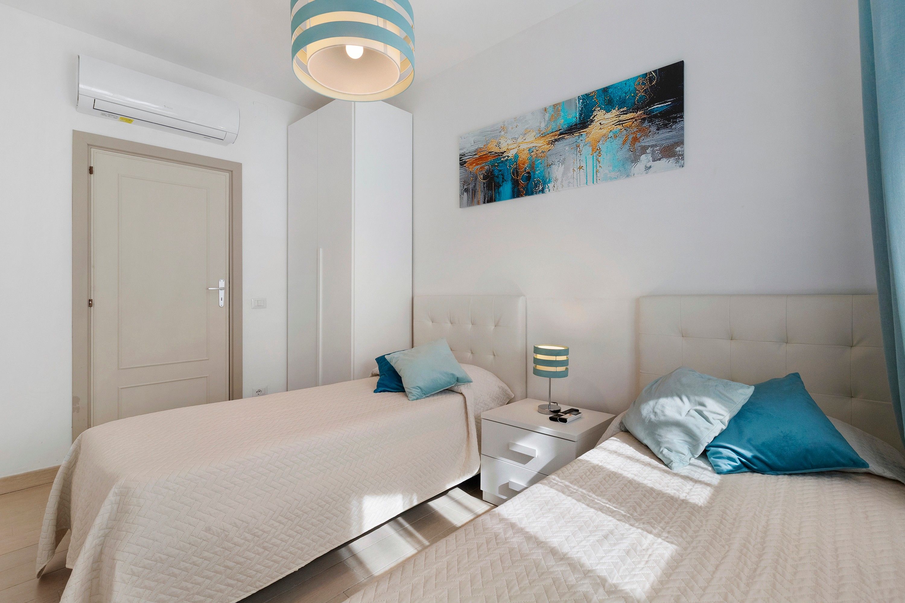 Une chambre d’hôtel avec deux lits, une table de chevet avec une lampe, un tableau au mur et un appareil de climatisation.