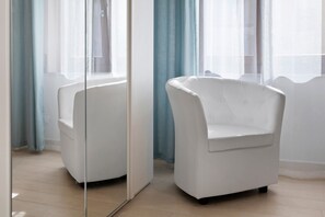 Una habitación con un sillón blanco, un separador de vidrio y vistas a través de una ventana con cortinas azules.