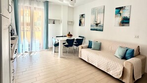 A modern living room with a sofa, dining table, chairs, and artwork on the walls. - Holiday Home 'Apartment For 6 People Near The Sea' with Balcony, Wi-Fi and Air Conditioning (Alghero)