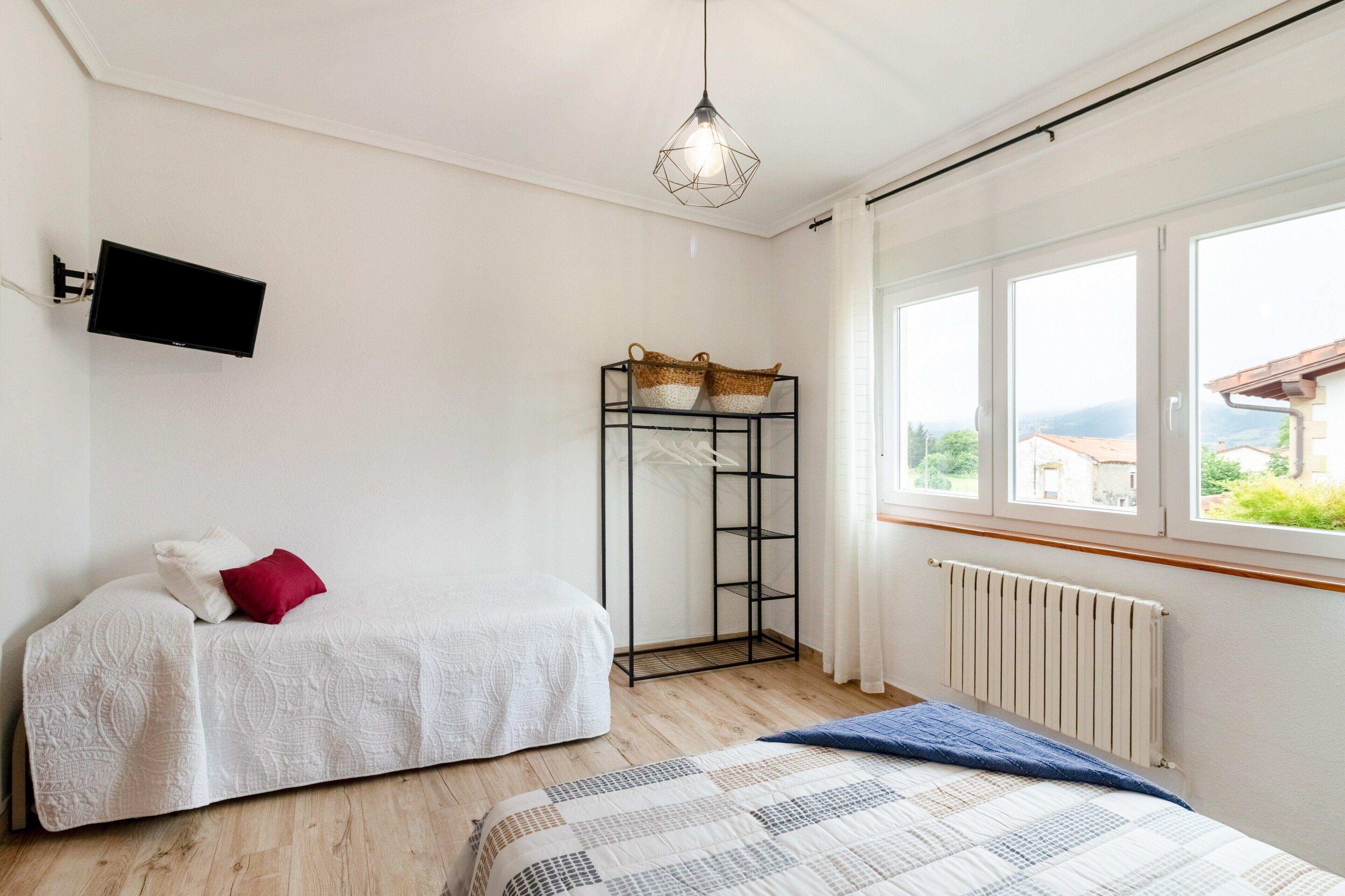 A bedroom with a bed, a small table with a basket, a radiator, and a window with a view.