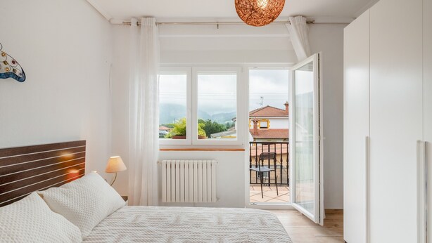 A bedroom with a bed, a radiator, a window, and a view of a balcony with a table and chairs.