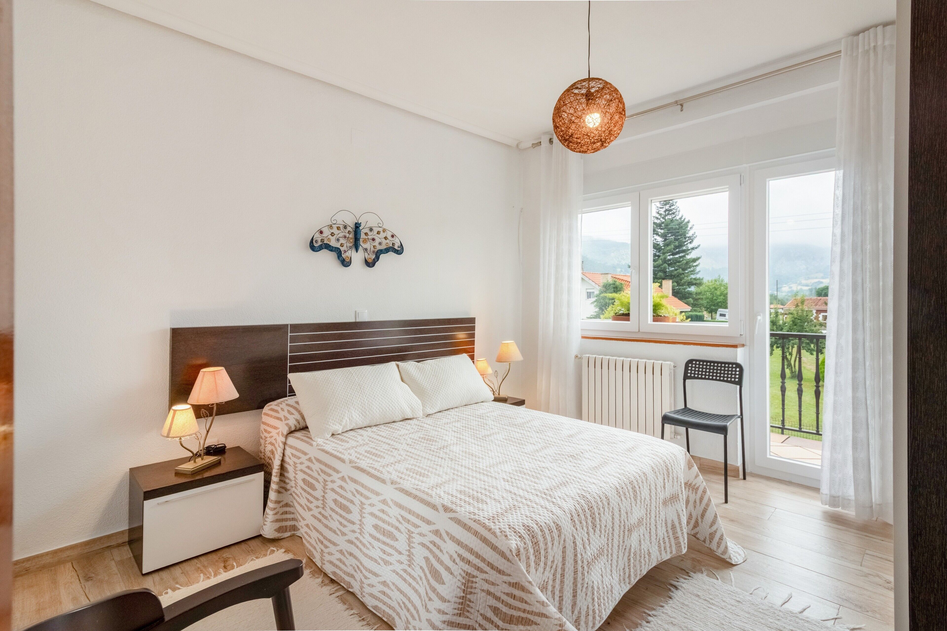 Quarto com cama, luminárias de cabeceira, mesa de cabeceira, uma cadeira, janela com vista e arte decorativa de borboleta na parede.
