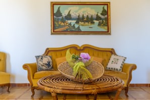 Ein Korbstuhl mit gewebtem Korb und einer violetten Blume, ein gelbes Sofa mit gemusterten Kissen und ein Landschaftsbild an der Wand.