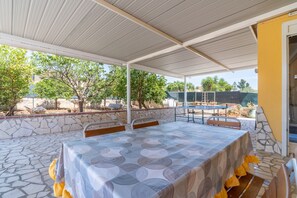 Een overdekte eethoek buiten, met stenen vloer, een tafel met tafelkleed met patroon en stoelen.