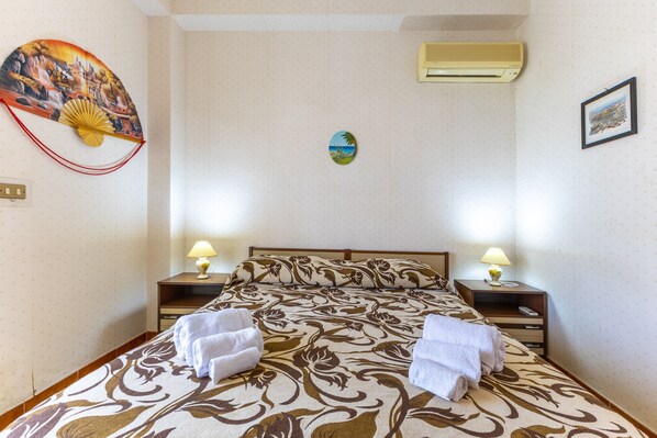 A hotel room with a bed, two bedside tables with lamps, a wall-mounted air conditioner, a decorative fan, and a small framed picture.
