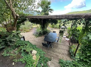 Een houten terras met een tafel en stoelen, omringd door weelderig groen en een pergola.