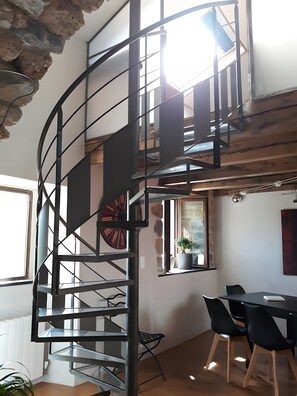 Een wenteltrap met metalen leuning, een eethoek met een tafel en stoelen, en een plant in een pot op een vensterbank.
