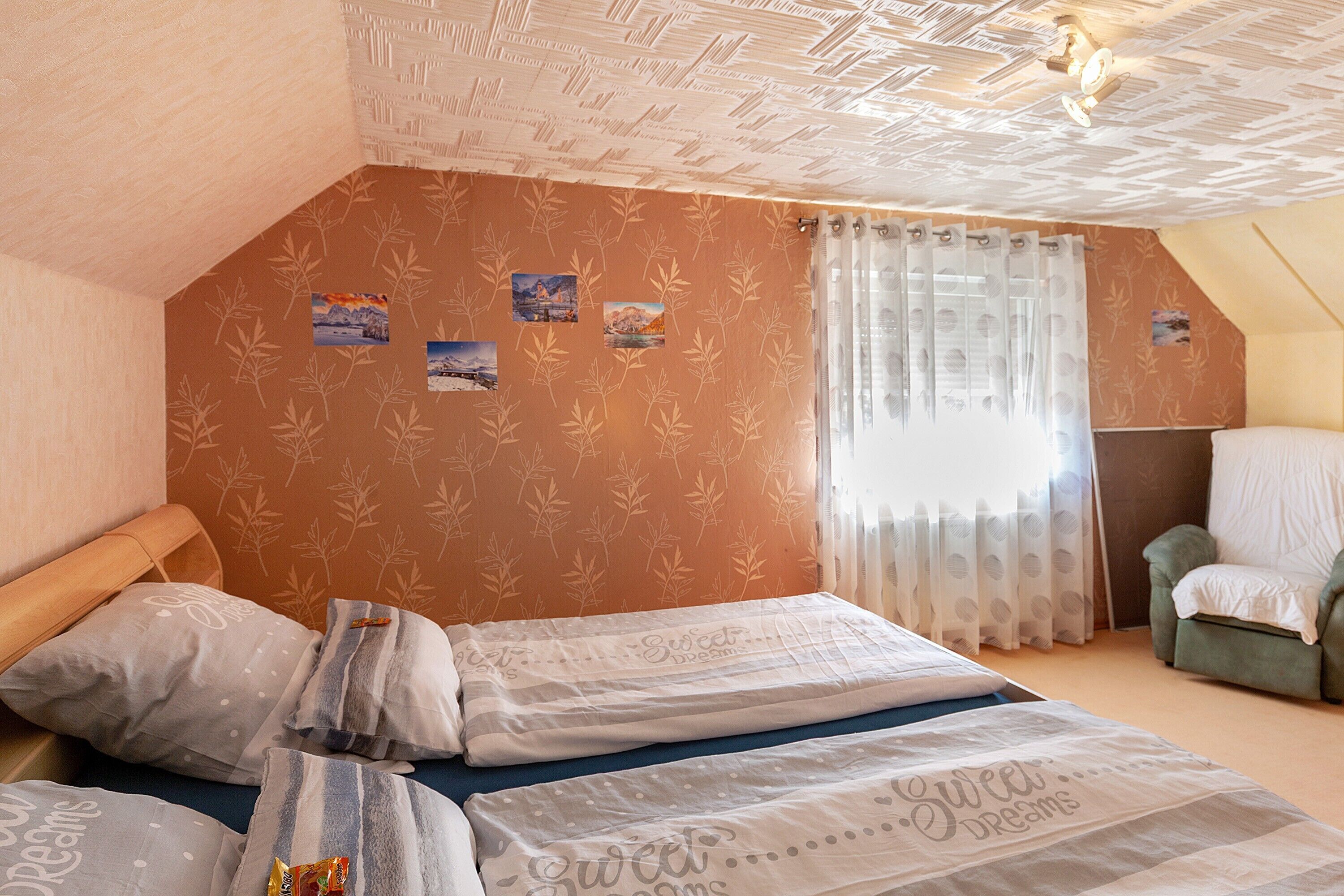 Ein Schlafzimmer mit schrägem Dach, einem Bett mit vielen Kissen, einem Sessel und einem Fenster mit transparenten Vorhängen.