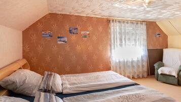 Ein Schlafzimmer mit schrägem Dach, einem Bett mit vielen Kissen, einem Sessel und einem Fenster mit transparenten Vorhängen.