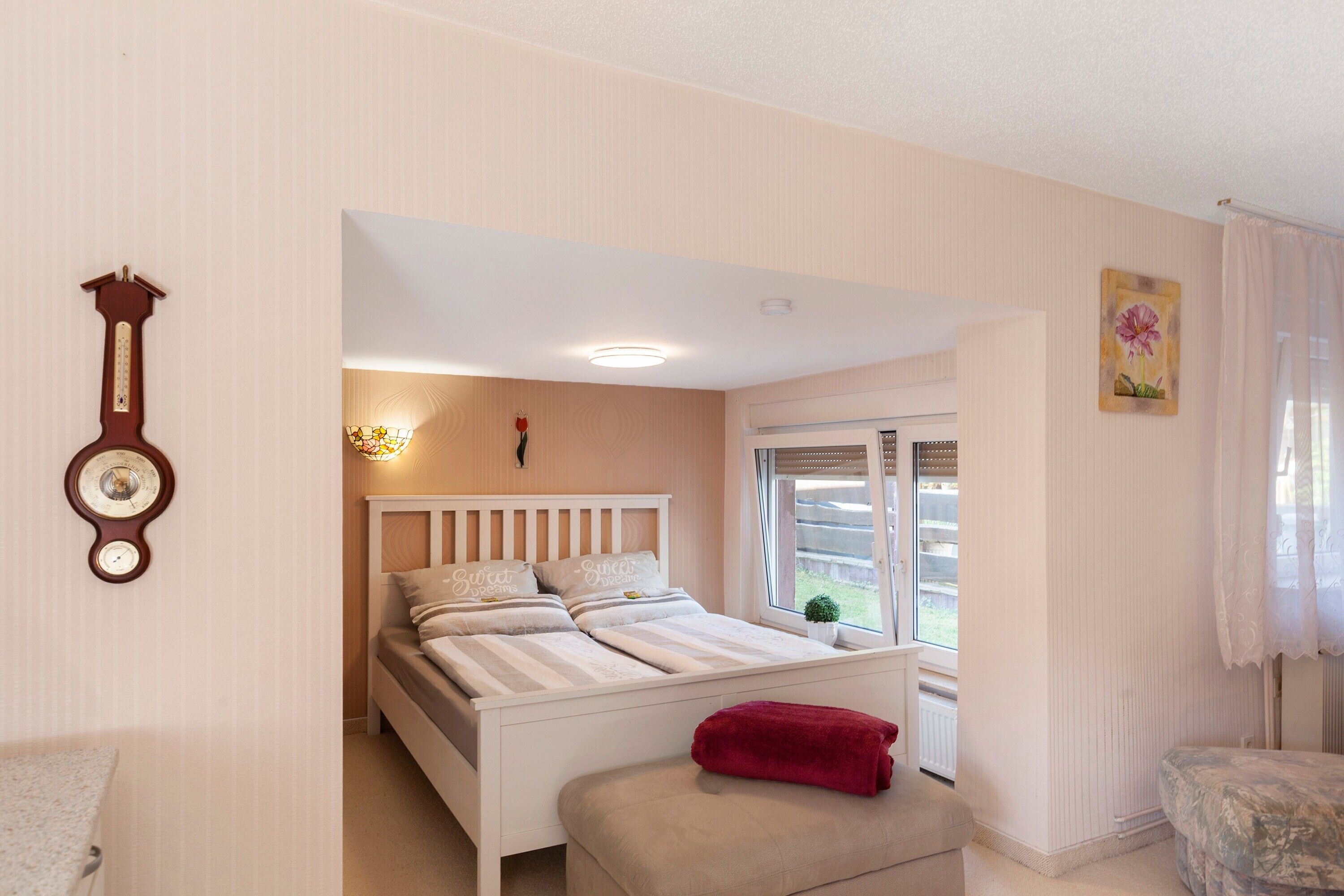Un dormitorio con cama, mesita de noche, un cojín rojo, un reloj de pared y ventana con vistas al exterior.